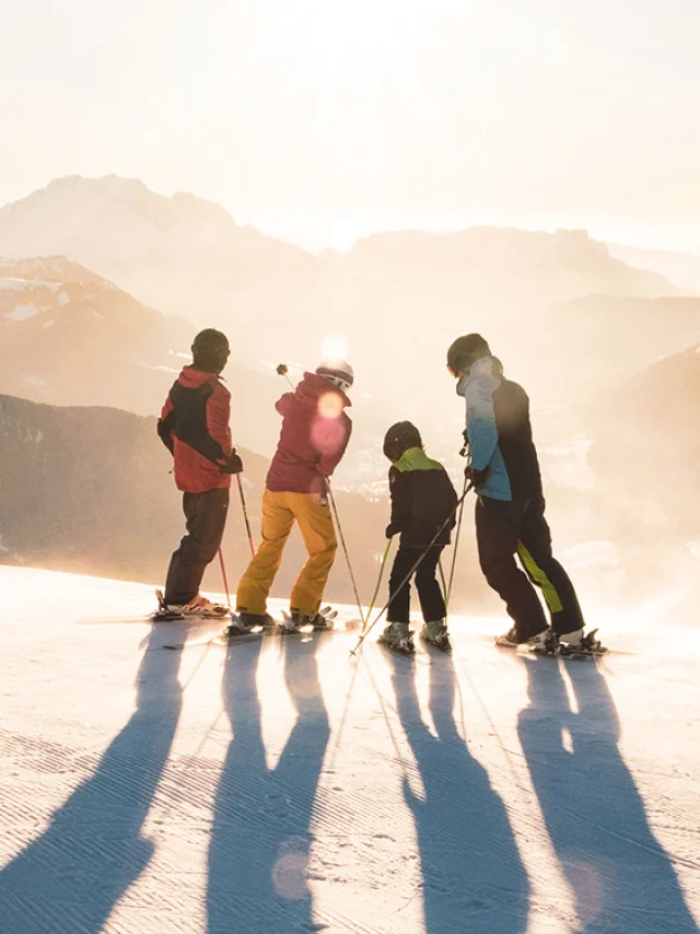 Eine Familie mit zwei Kindern steht auf Skiern am oberen Ende einer Piste und betrachtet den Sonnenuntergang mit Blick auf die Aravis-Kette. Einer hat einen blau-schwarz-grünen Skisatz, ein anderer einen rot-gelben, ein weiterer einen grün-schwarzen und der letzte einen rot-schwarzen. Einer der Skifahrer in der Mitte zeigt mit seinem linken Stock auf den Gipfel eines Berges. Die Atmosphäre ist sehr mondähnlich mit weißem/goldenem Licht. Es ist ein schöner Abschluss eines Skitages.